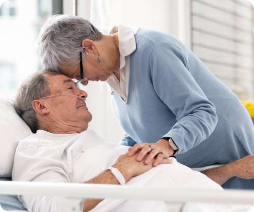 Caregiver gently comforting an older adult resting in bed at home.

